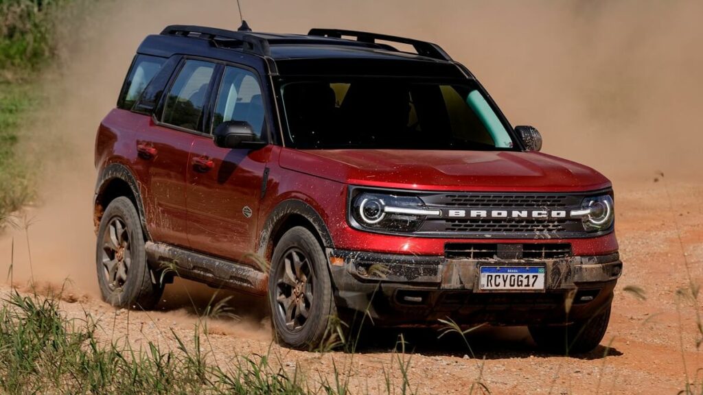 Lateral frontal de uma Ford Bronco 2023  na cor vermelha com partes em preto rodando por uma estrada de barro