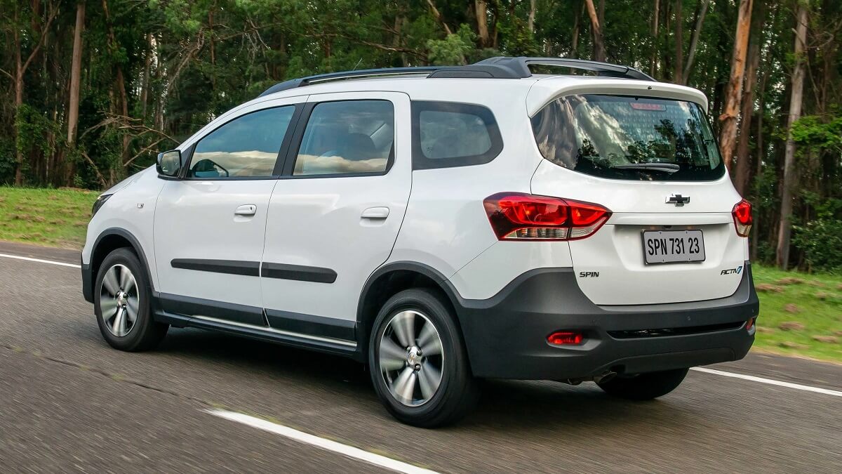 Lateral traseira de uma Chevrolet Spin 2023 na cor brancas, com partes em preto rodando pelo asfalto com vegetação ao fundo.