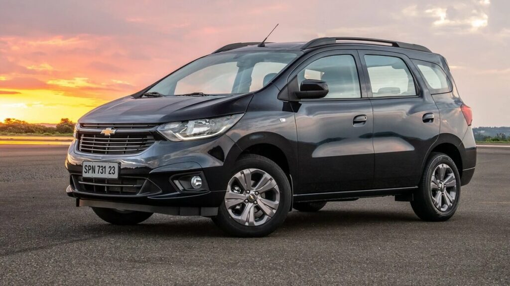 Lateral frontal de uma Chevrolet Spin 2023 na cor preta, estacionada em um asfalto durante um por do sol.