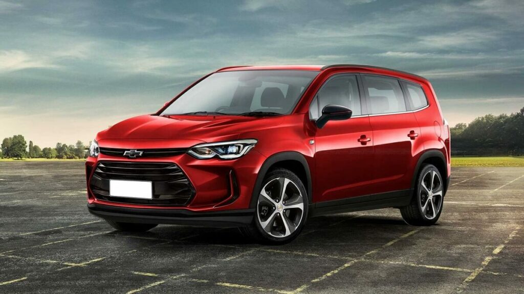 Lateral frontal de um Chevrolet Spin 2023 na cor vermelha, com partes em preto estacionado em uma pista com um gramado ao fundo