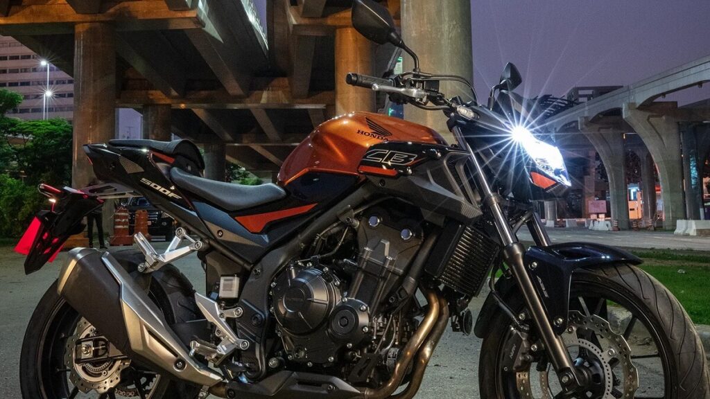Lateral de uma CB 500F 2023 laranja, com partes pretas abaixo de uma ponte em uma noite.