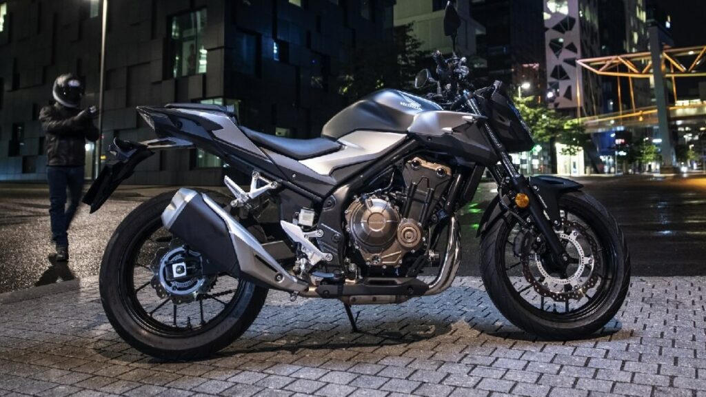 Lateral de uma CB 500F 2023 na cor prata, com partes em preto, estacionada em uma calçada a noite.