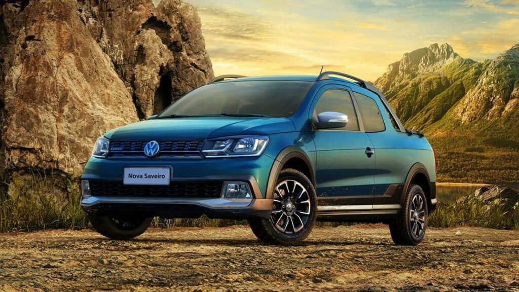 Lateral frontal de uma VW Saveiro 2023 na cor azul, estacionada em um chão de barro com uma paisagem montanhosa.