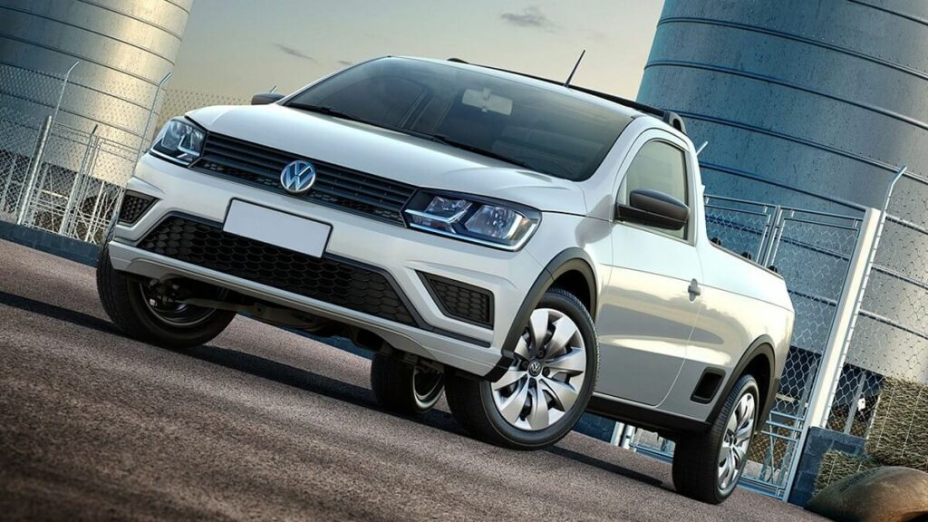 Lateral frontal de uma VW Saveiro 2023 na cor branca, estacionada em um asfalto com um reservatório ao fundo.