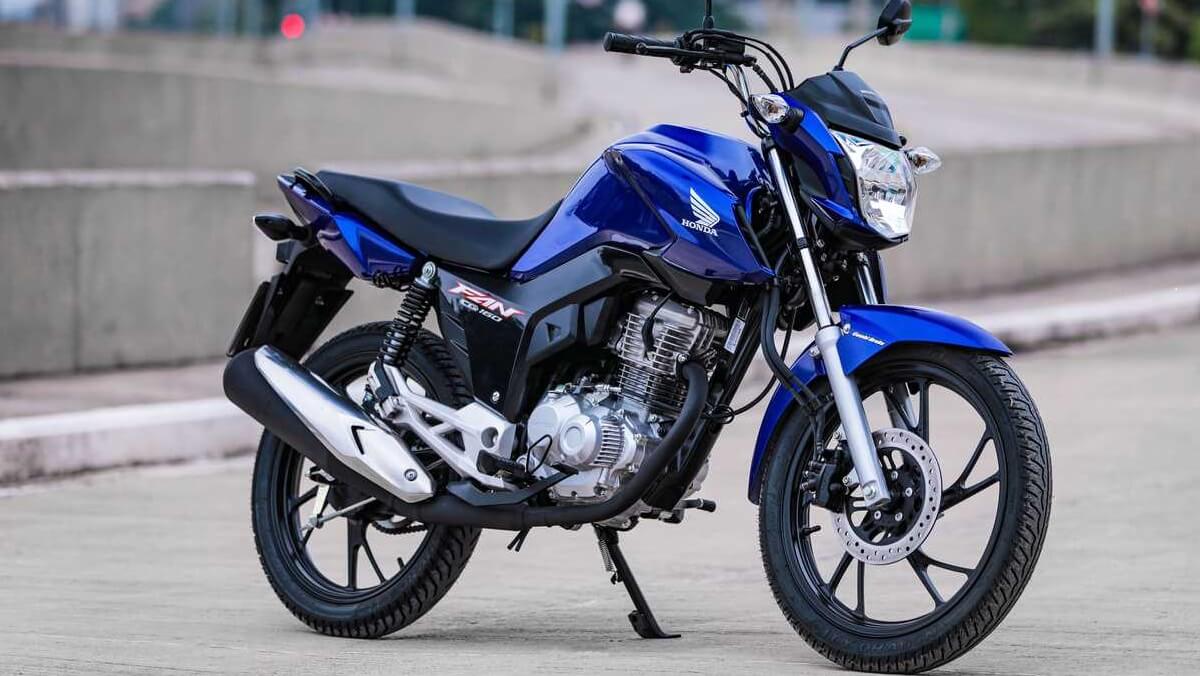 Foto frontal da Honda CG 160 Fan 2023 na cor Azul Perolizado a moto se encontra em uma rua próxima da rodovia
