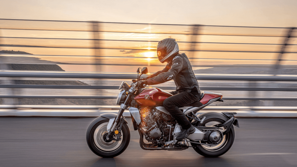 Foto lateral de uma Honda CB 1000R 2023 vermelha andando em uma estrada com por do sol de fundo