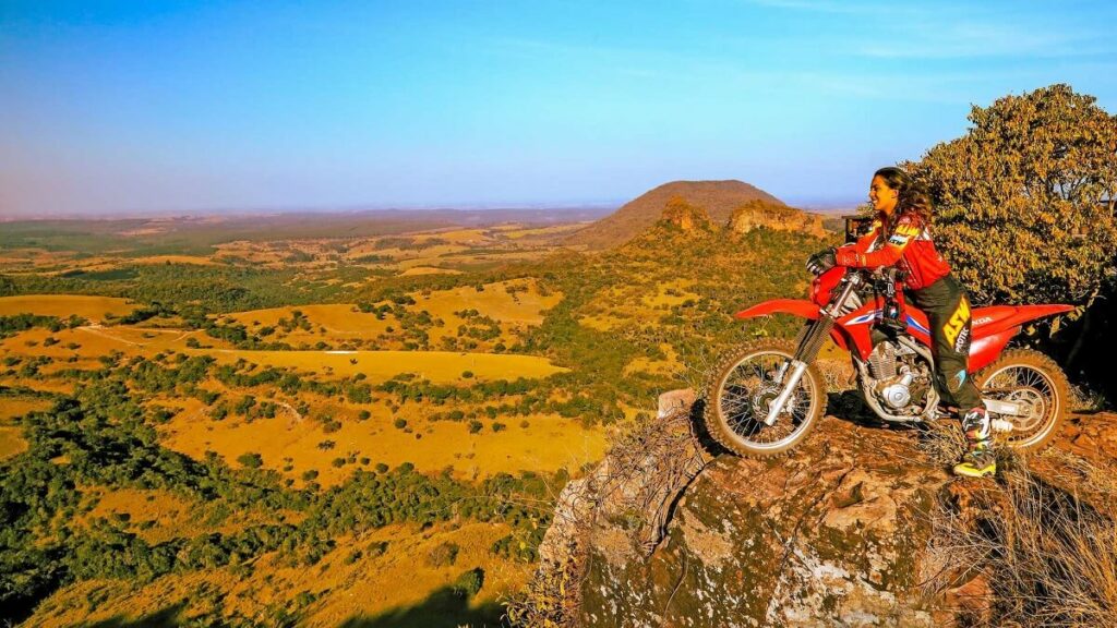 Foto de uma pessoa em cima de uma pedra em um morro, a mulher esta montada na moto olhando para a bela paisagem, a moto é uma Honda CRF 250 F 2023
