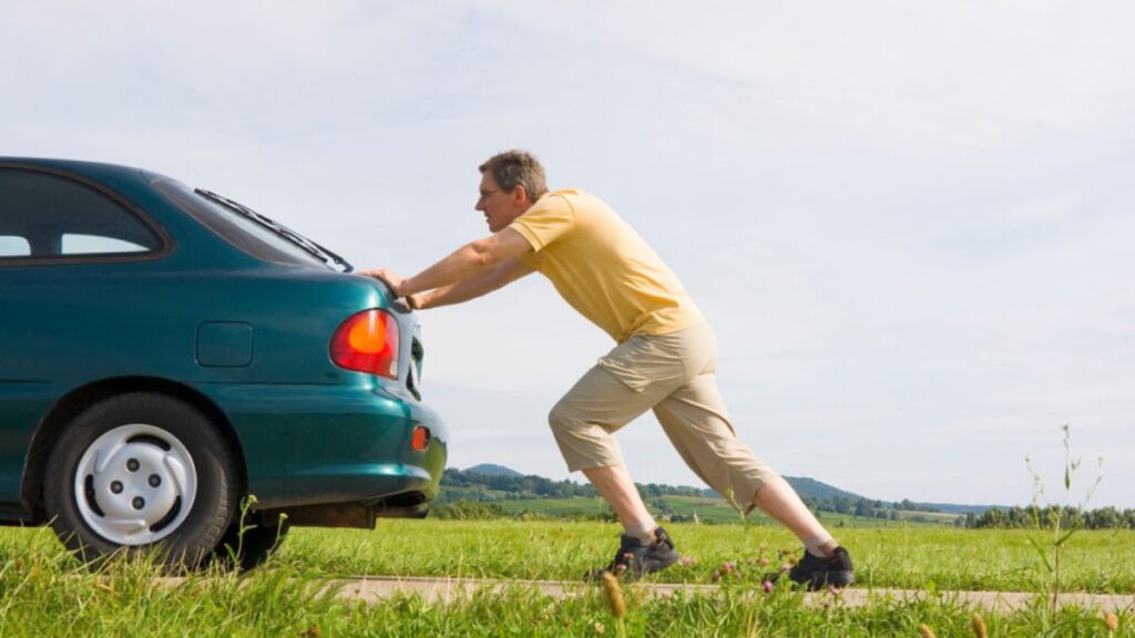Homem empurrando carro verde na estrada.