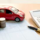 Um mini carro e 5 mmontes de moedas em cima de um papel do lado de uma calculadora e uma caneta, isso significando os juros que estão altos.