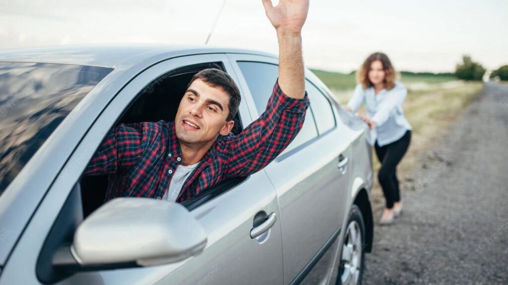 Um homem com a mão levantada dento de um carro prata com uma mulher empurrando.