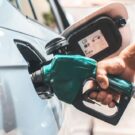 Pessoa segurando um pistola verde de uma bomba de combustível abastecendo um carro cinza.