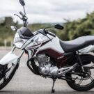 Honda CG 150 parada na diagonal branca.