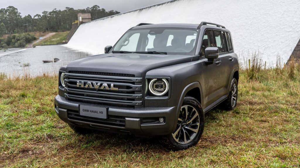 GWM Haval H9 2026 cinza parado na diagonal em um gramado com uma cachoeira no fundo.