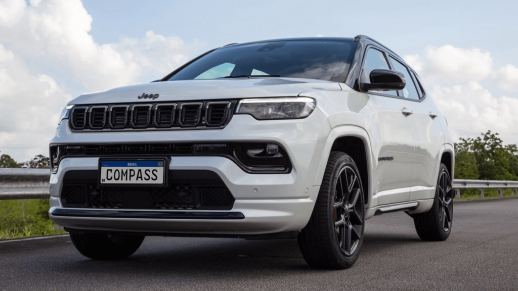 Jeep Compass Sport 2026 branco em movimento em um asfalto com matos verdes na beirada e um céu azul cheio de nuvens.