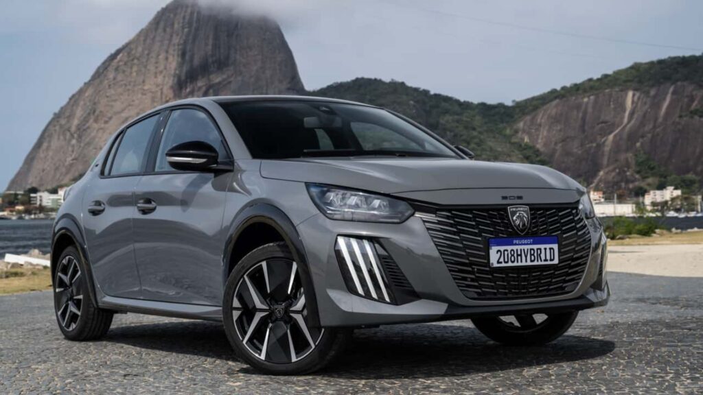 Peugeot 208 2026 híbrido cinza parado na diagonal com o Pão de açúcar no fundo.
