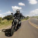 Homem de preto em cima de uma moto em velocidade na estrada de asfalto de dia.