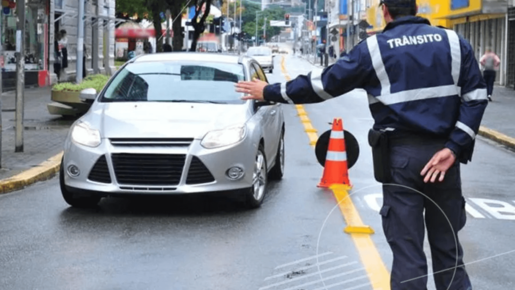 Guarda de trânsito com a mão estendida para o carro prata que se aproxima.