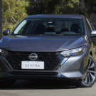 Frente do Nissan Sentra Advance 2025 cinza parado na diagonal com os faróis acesos.