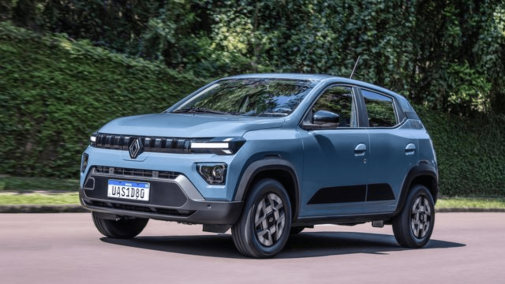 Renault Kwid E-Tech 2026 azul em movimento na estrada com os faróis acesos e um muro com plantas no fundo.