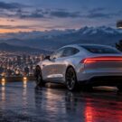 Carro elétrico branco ao lado de um carregador, com vista para uma cidade e montanhas ao pôr do sol